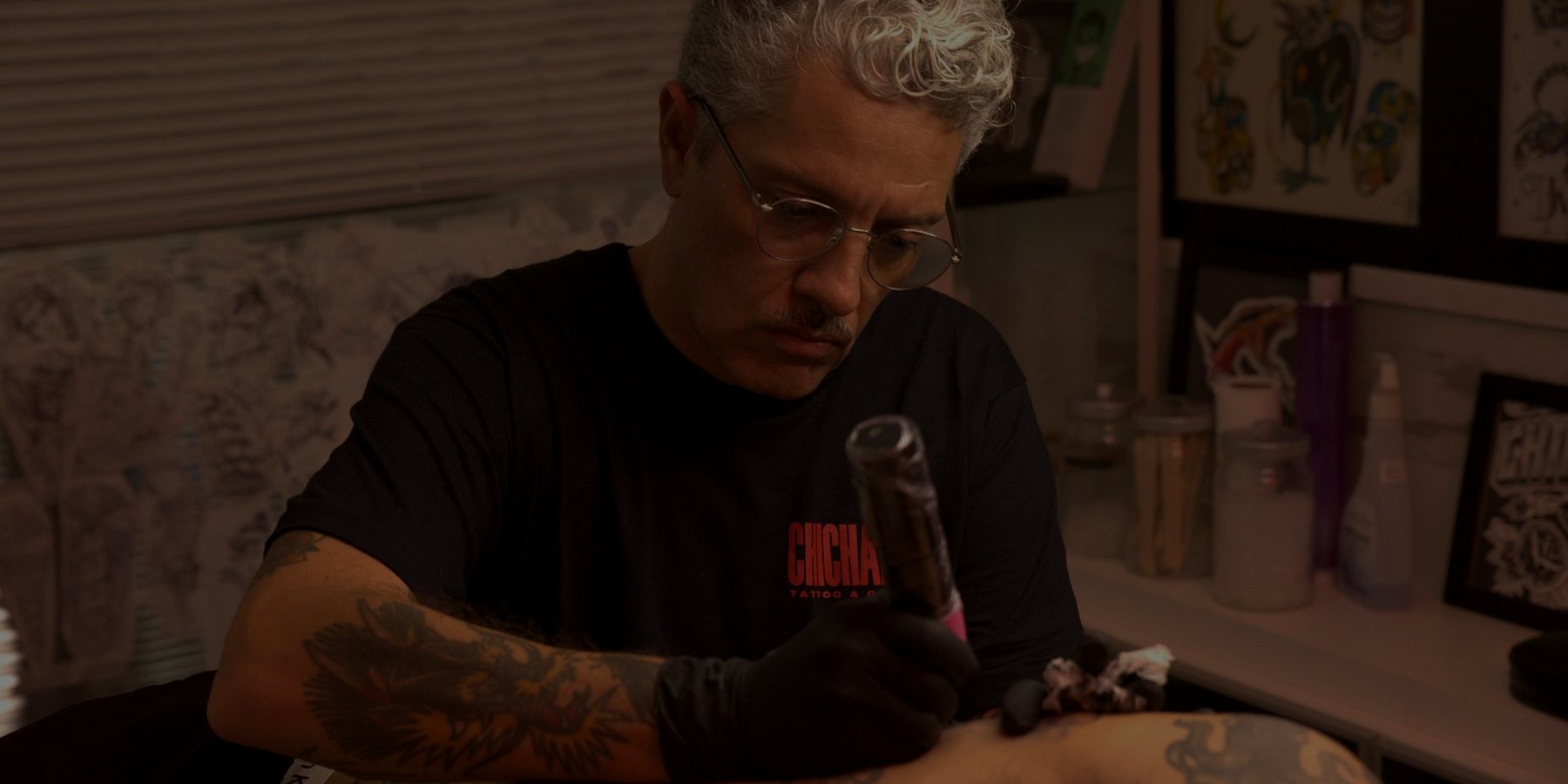 Diego Mena tatuando en el estudio, con las láminas al fondo